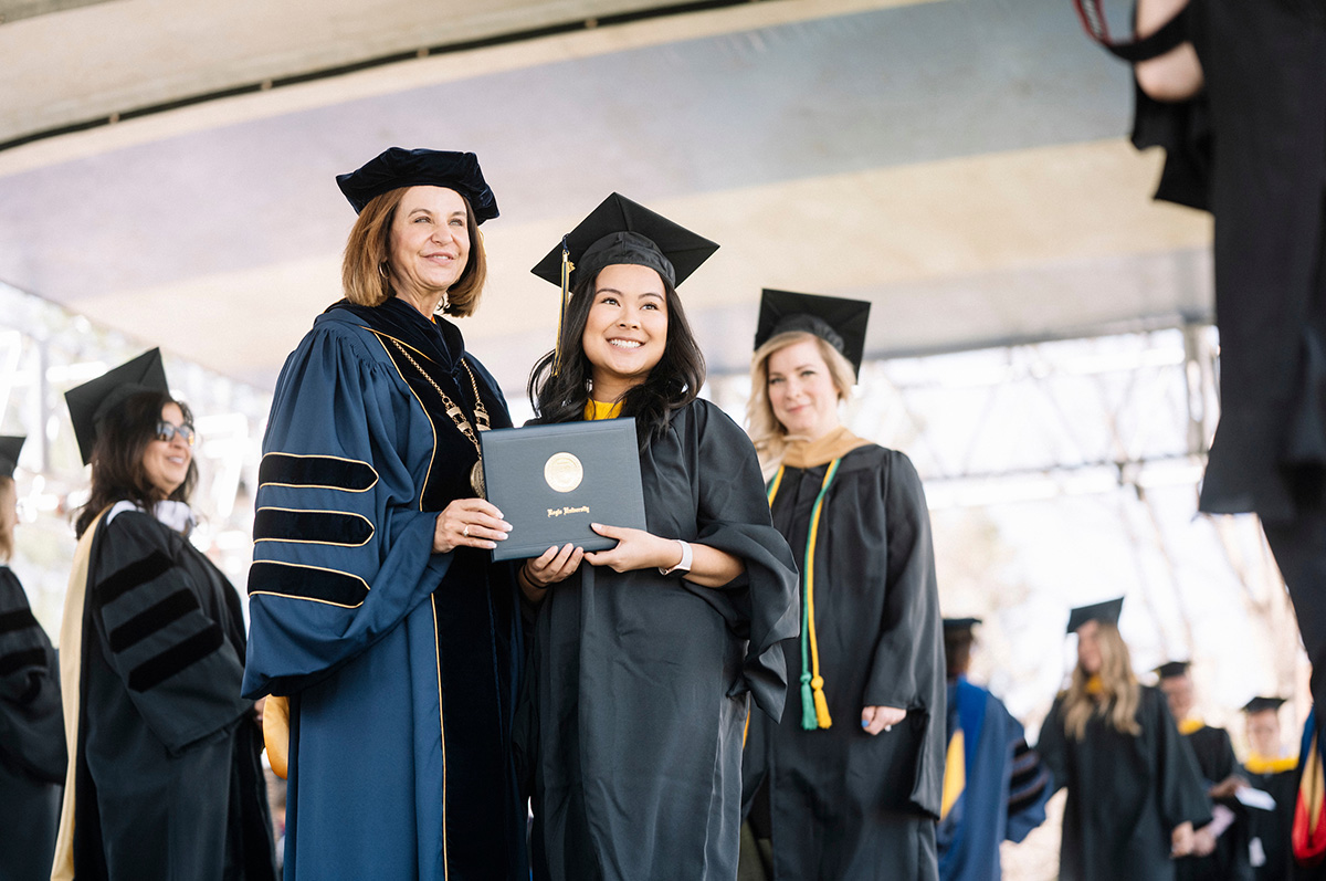 A smiling graduate in cap and gown shakes hands with faculty as she accepts her diploma