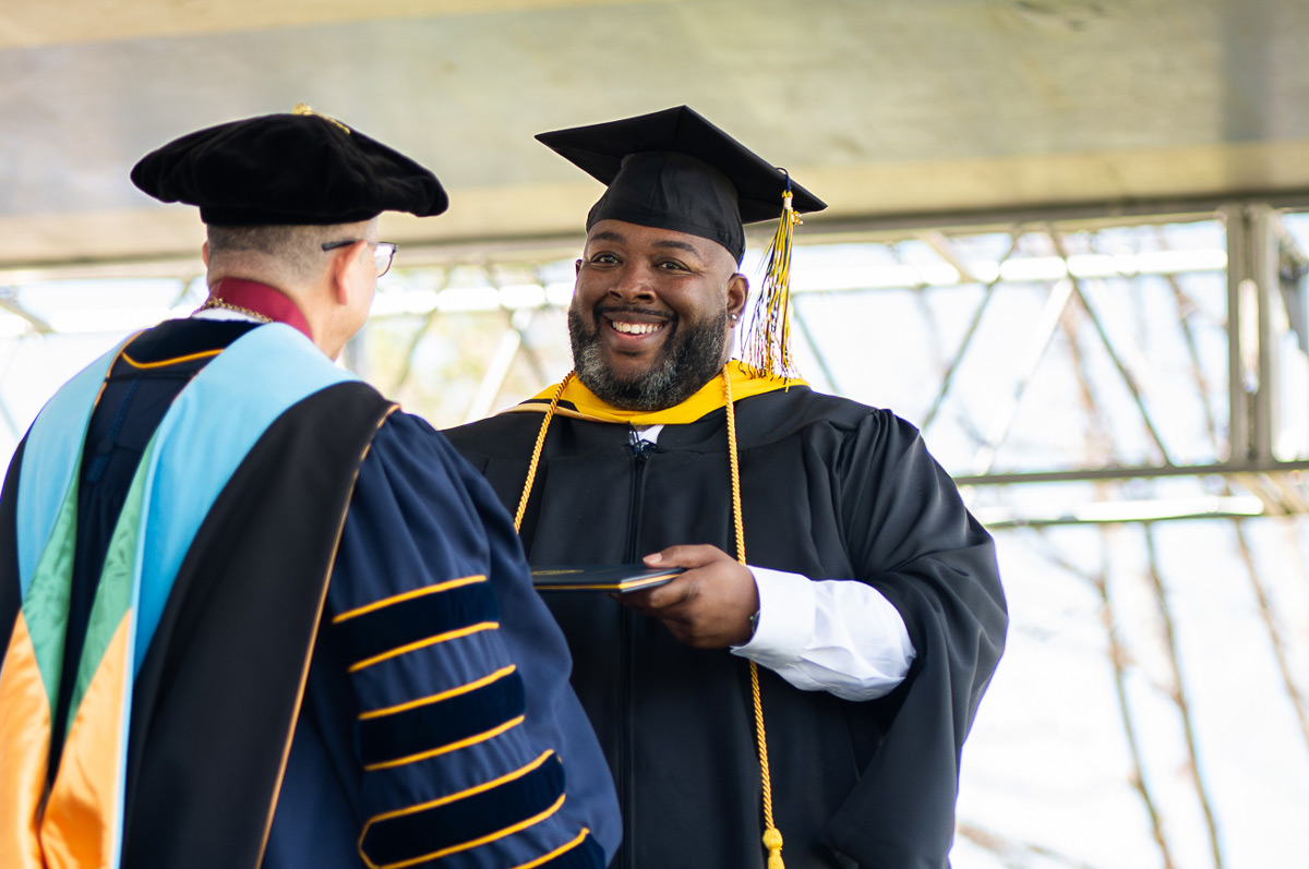 A smiling graduate in cap and gown receives a diploma from a faculty member in academic regalia on stage