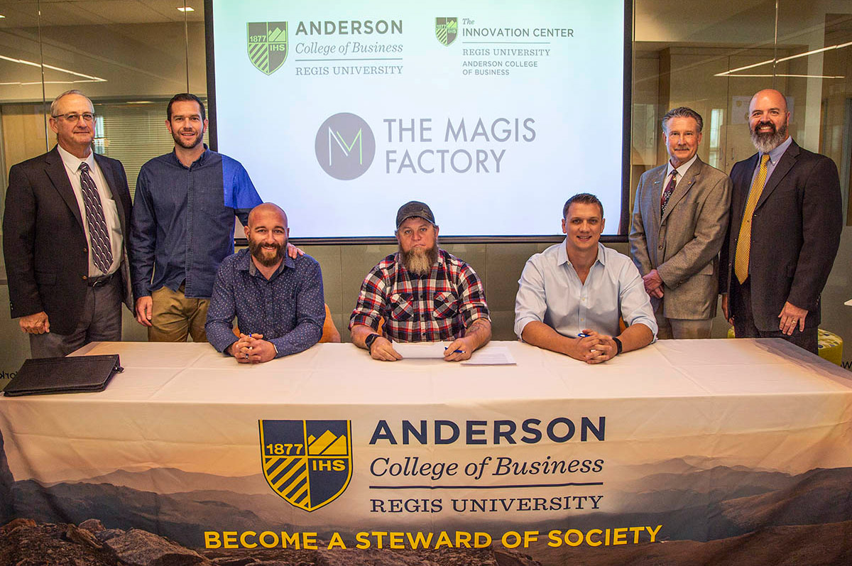 three men sit at a table at Regis surrounded by their faculty mentors 