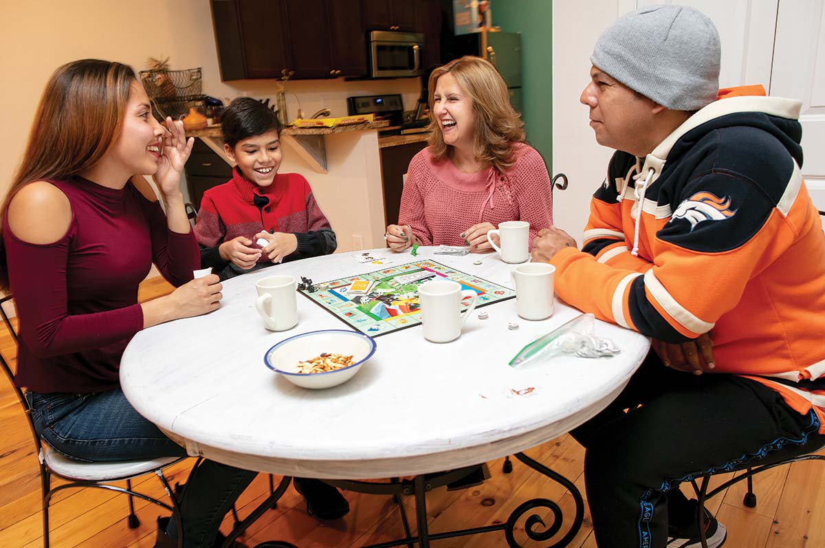 The Nuñez-Pulgar family and Ludy Yevara sit at a table in an apartment playing Monopoly, laughing and enjoying snacks.