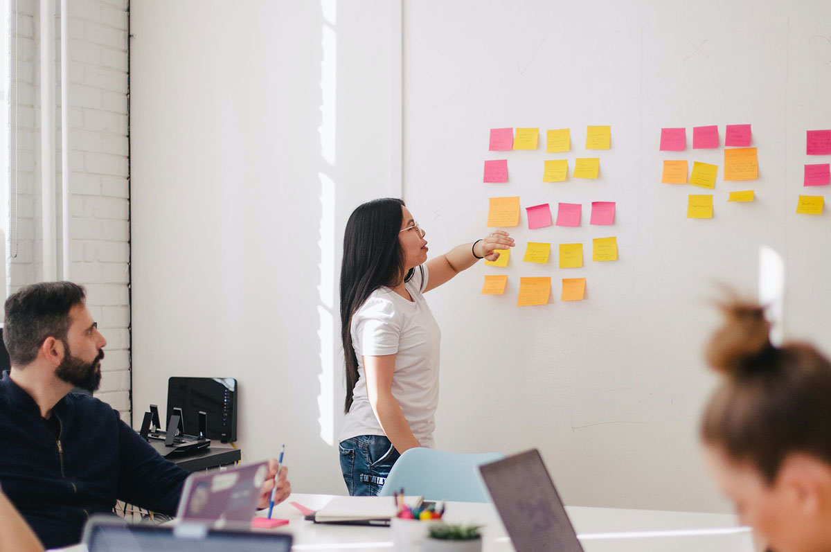 men and women sit at a conference table in an office while a woman stands in front of a white board and gestures at assorted sticky notes