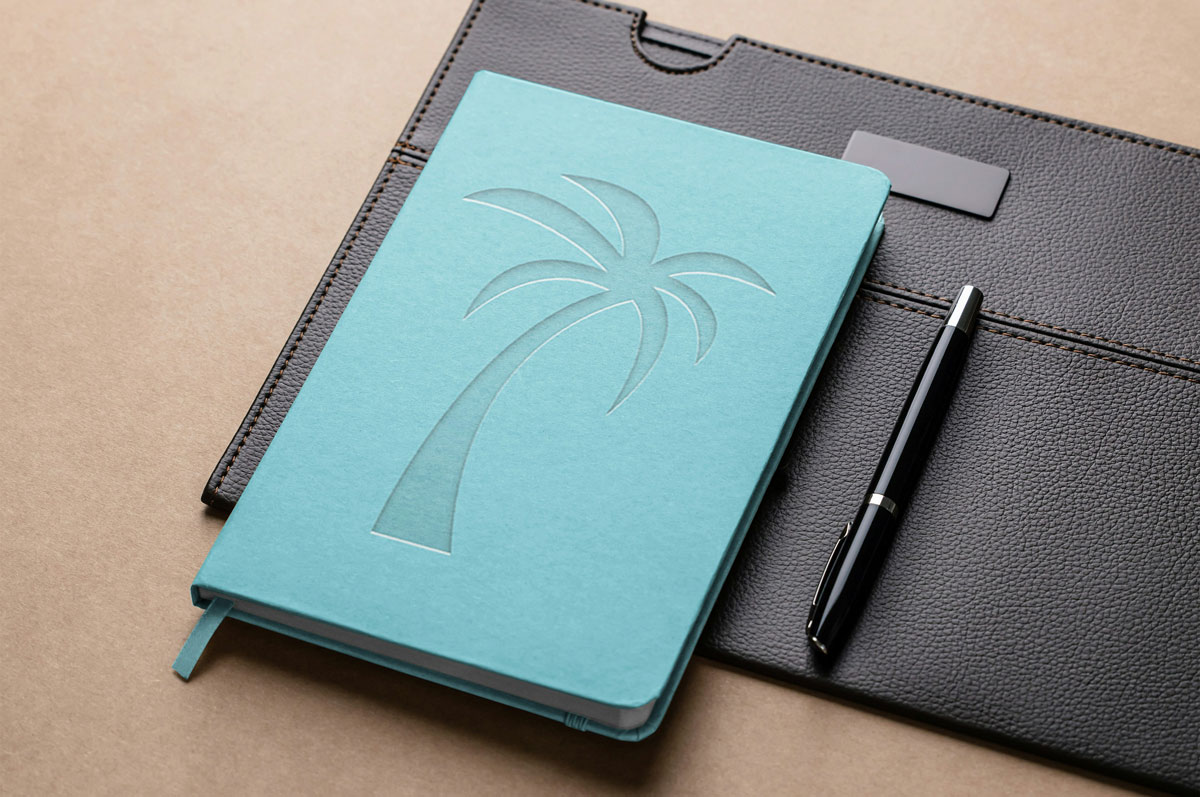 Turquoise notebook with an embossed palm tree resting on a dark brown leather folder, with a black pen beside it on a tan surface.