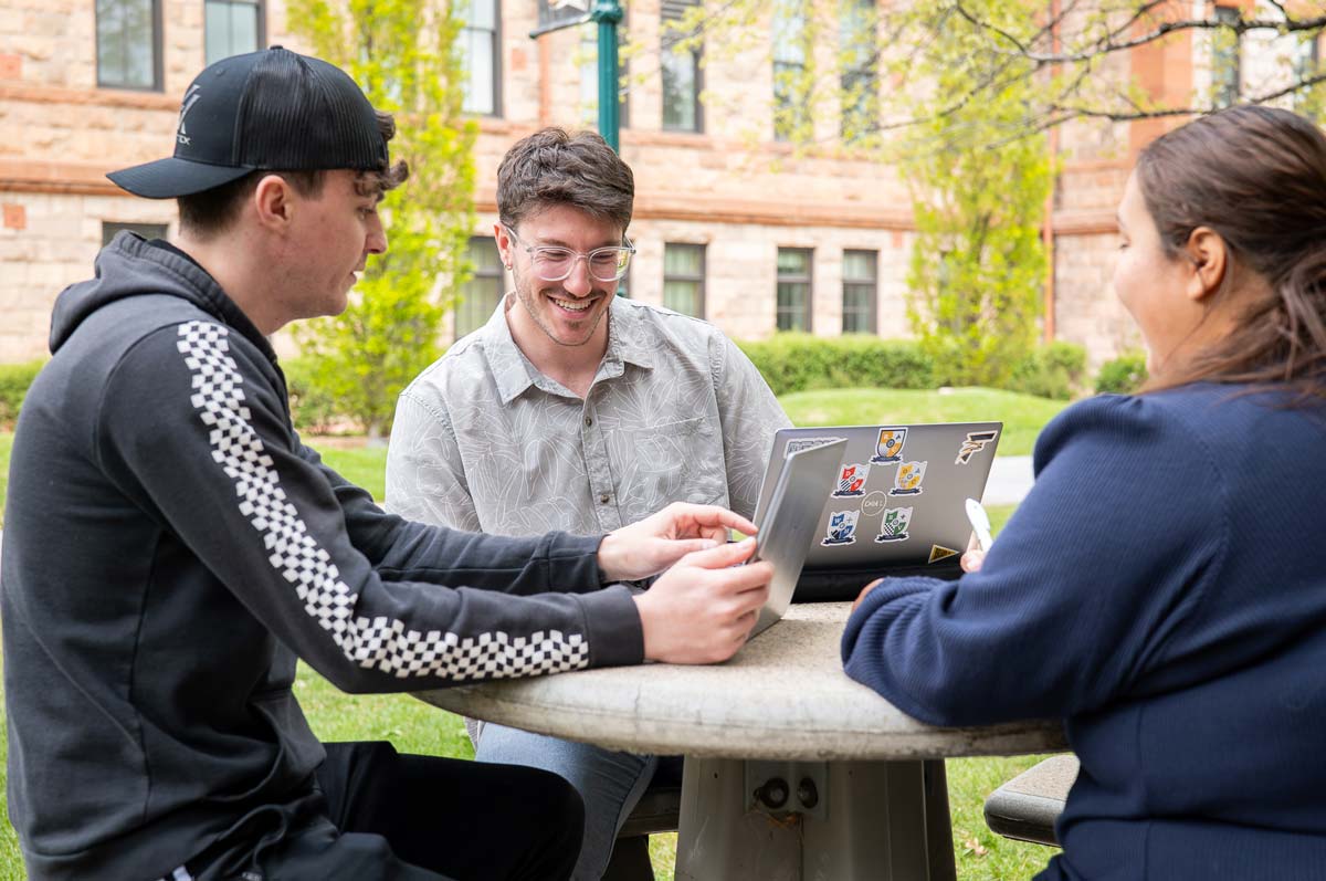 A group of three sit around a round table outdoors with laptops open and looking down at their work while smiling