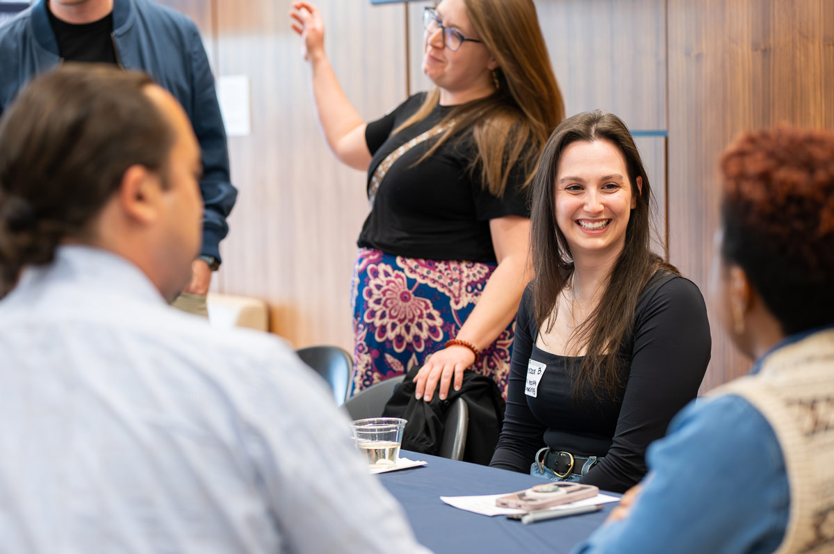 A small group of adult students sit around a table at a networking-style event, smiling and chatting. One woman in the foreground smiles warmly at others seated nearby, while another person stands behind gesturing as she speaks.