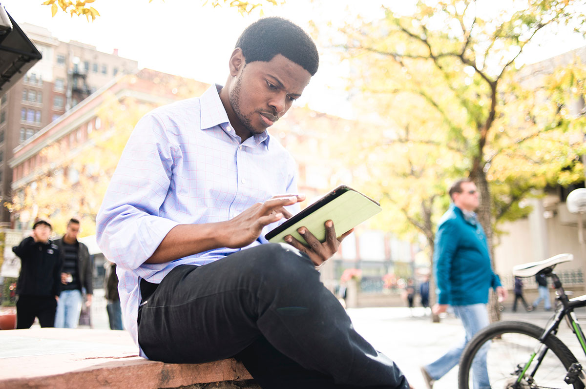 A young adult student dressed in business casual attire uses a tablet computer while seated on a bench in downtown Denver.