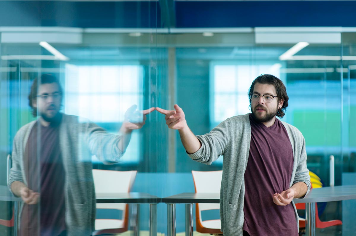 A young adult student points at a clear dry-erase board while giving a presentation, his reflection seen in the glass board. 