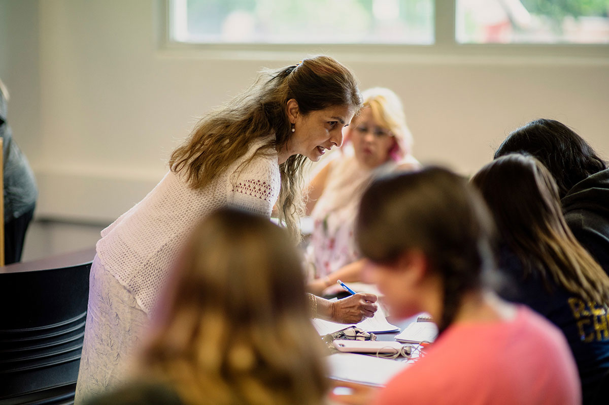 A woman professor holds a pen and leans slightly over a desk to speak with a group of young women students in a classroom. 