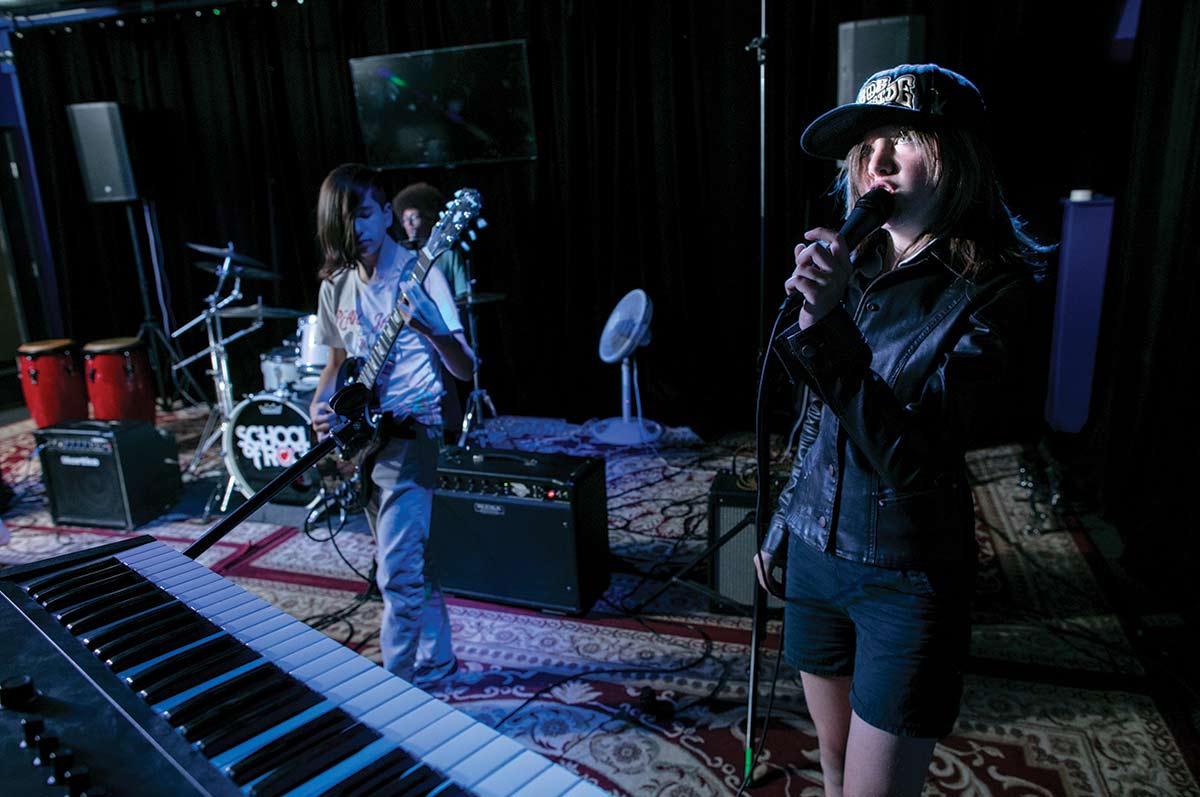 Three School of Rock students on stage. Student on the right is singing into a microphone. Student on the left is playing the guitar. Student behind them is on the drums.