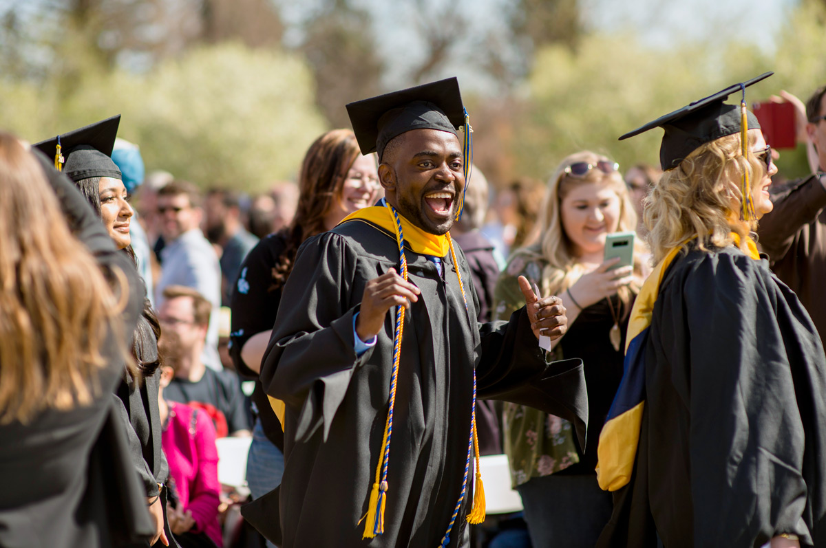 Graduates in caps and gowns walk outdoors among a crowd of people, one graduate in the center smiles and gestures excitedly