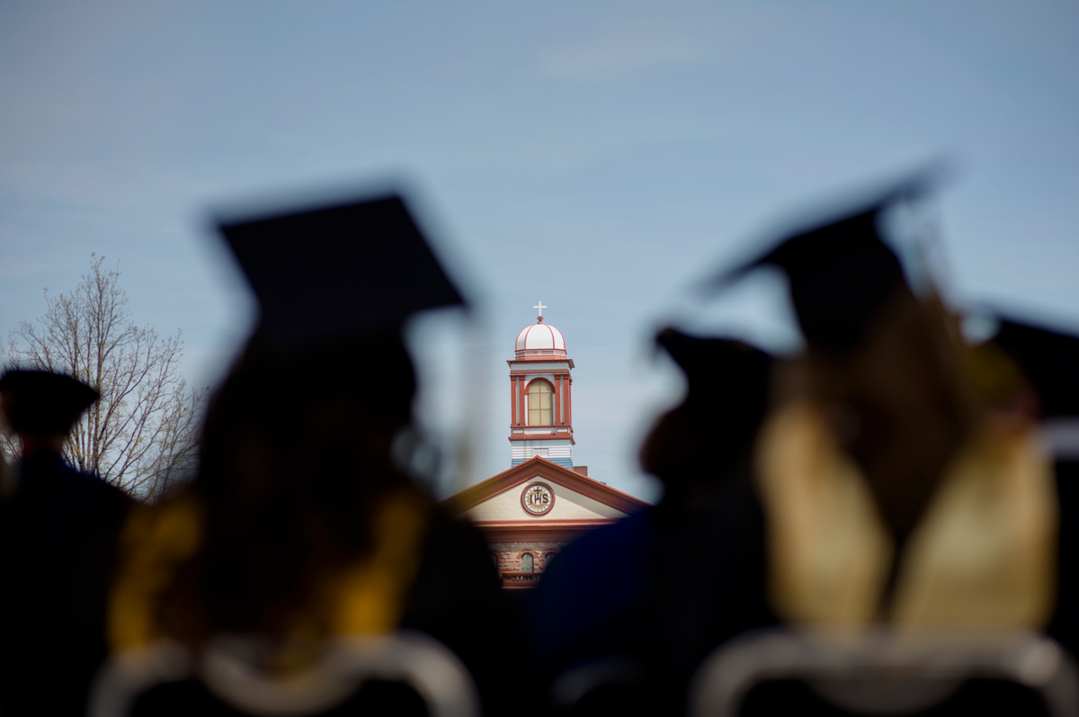 Graduates in caps and gowns sit facing Main Hall with the dome and cross in focus under a clear blue sky