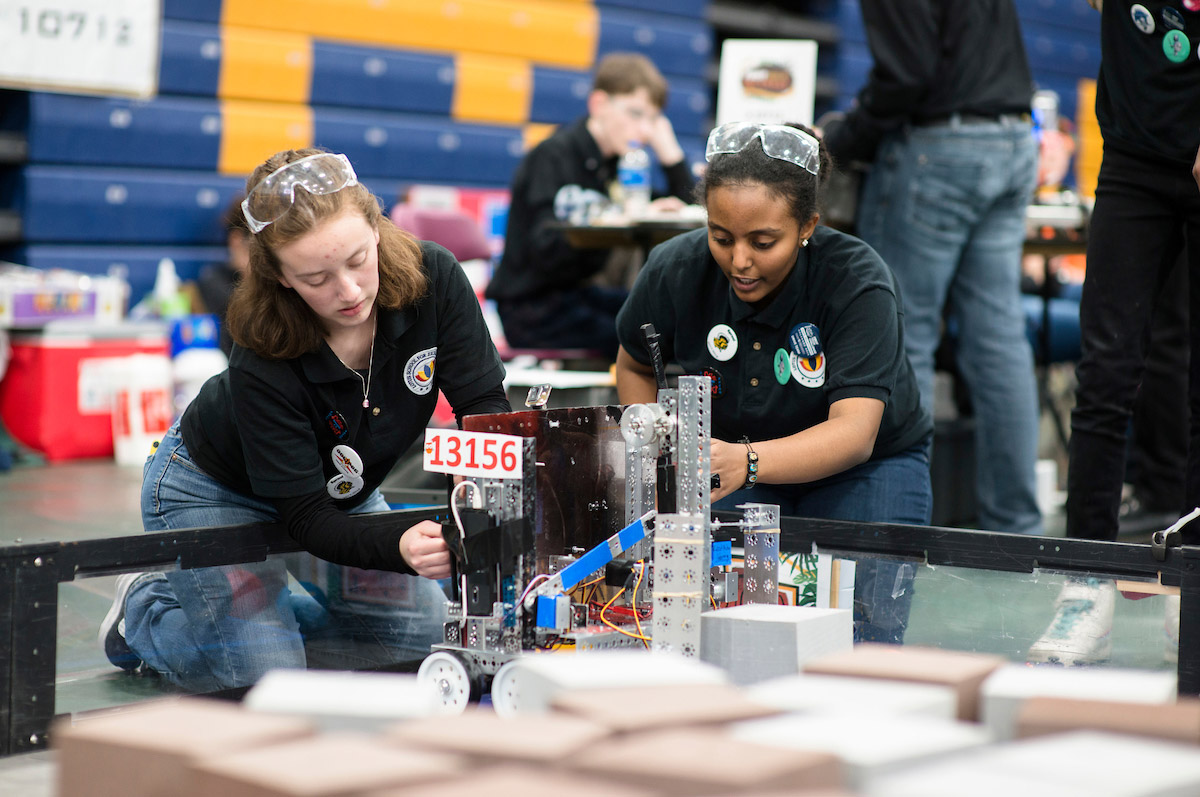 two young competitors kneel on the ground working intently on a small robot with wheels at the robotics competition