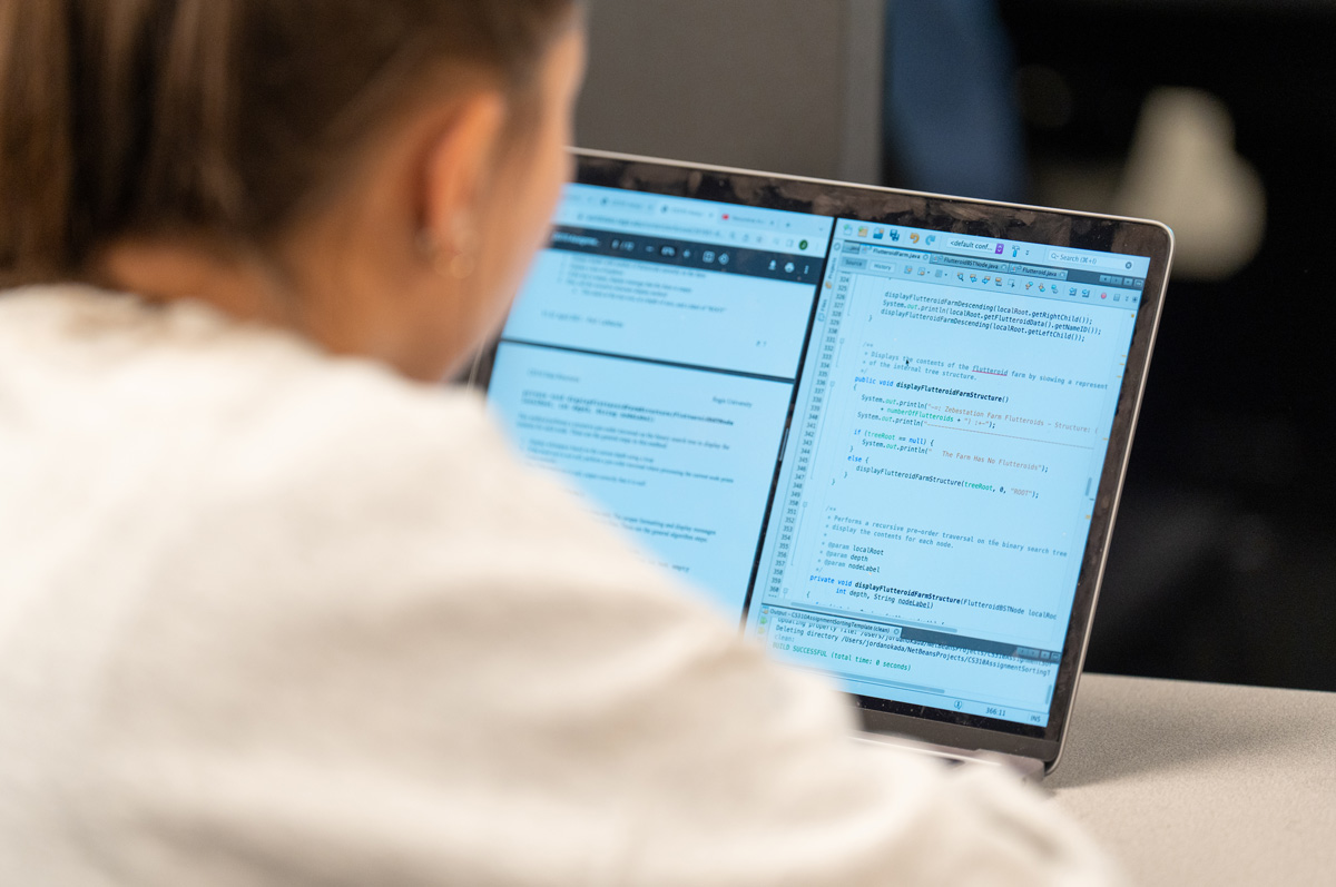 a student in a white sweater focuses intently on a laptop screen displaying programming code and text documents