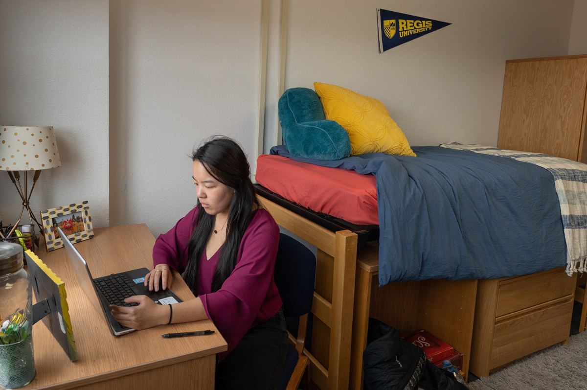 A student in a dorm room works on a laptop at a desk. The room features a neatly made bed with blue and red bedding and a Regis University pennant.