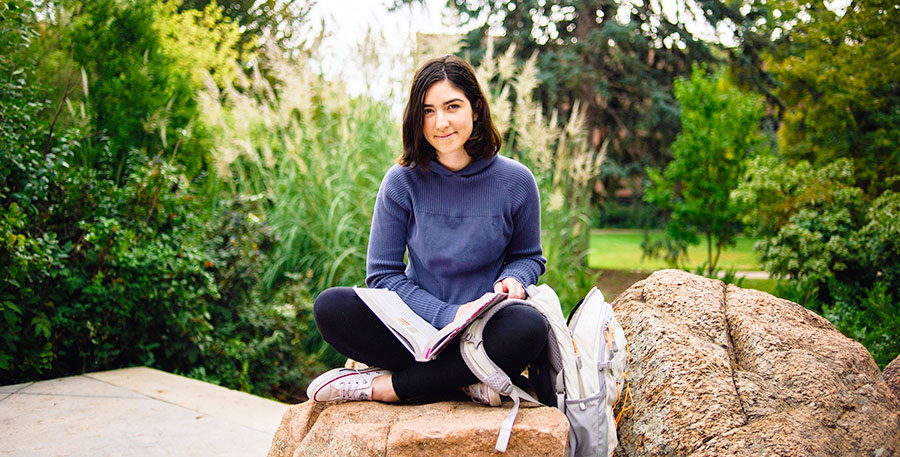 a student sits on a boulder reading from a textbook outdoors on the Northwest Denver campus