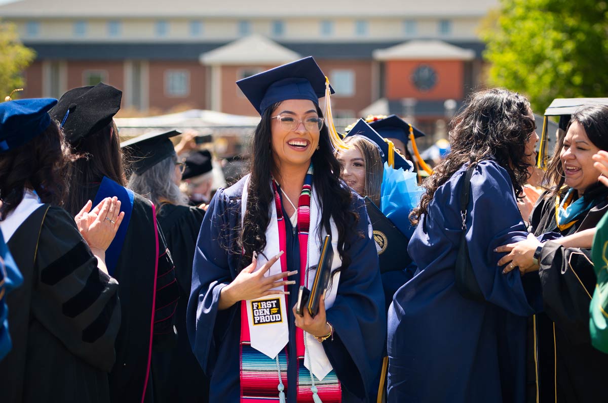Student smiles with their diploma and phone in left hand surrounded by other graduates and parents