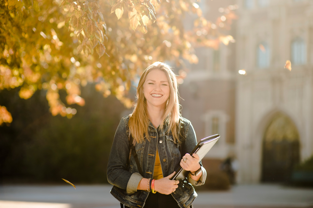 smiling young student in a denim jacket stands outdoors holding books, with golden autumn leaves and Carroll Hall in the sunlit background