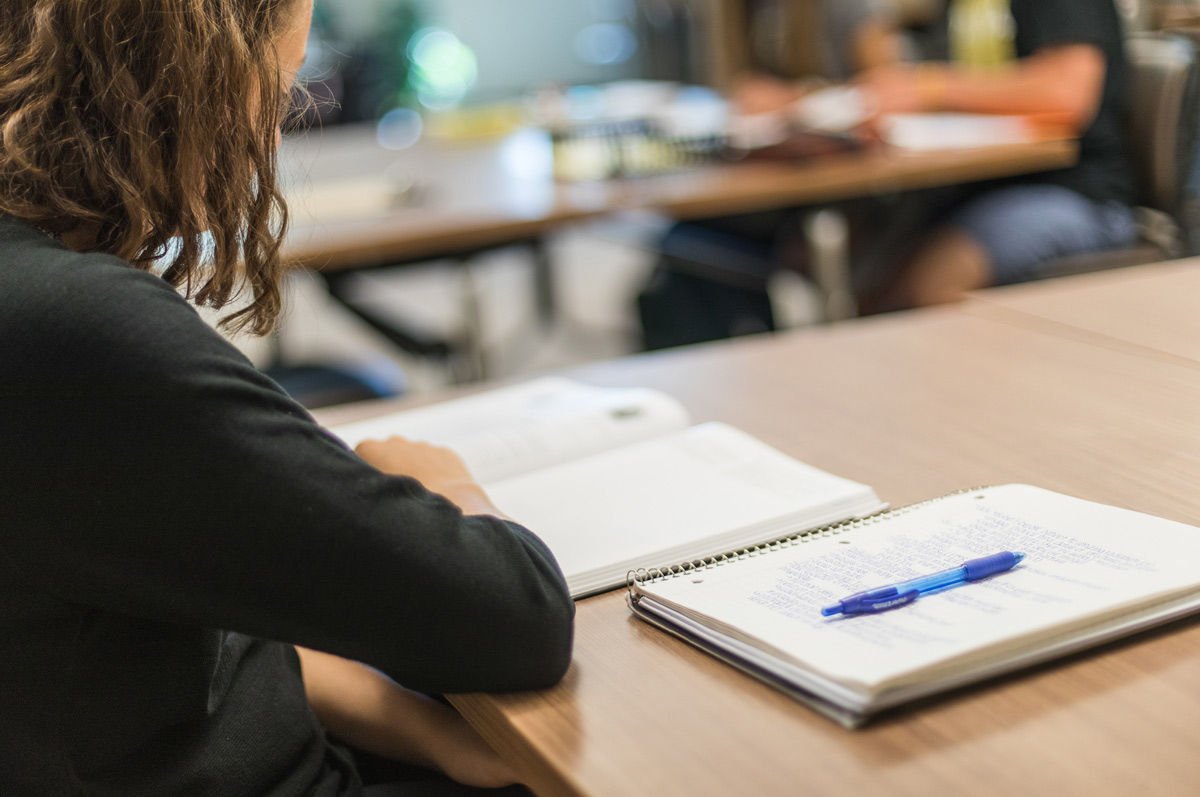 a student studies at a wooden table, focusing on an open book, with a notebook and a blue pen lying nearby