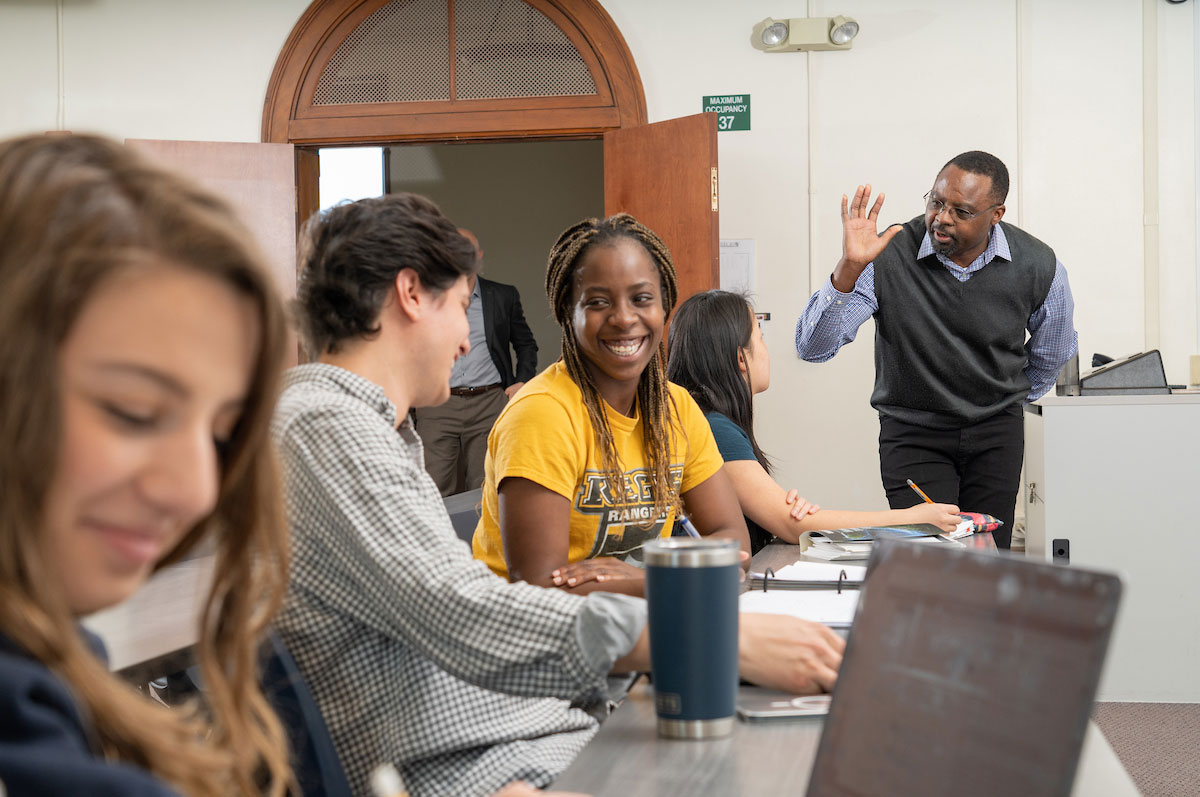 students in a classroom sit at desks with laptops talking and smiling while professor stands at the front of the room talking to student and gesturing broadly