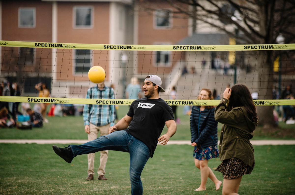 Regis students playing volleyball on campus lawn with the library building in background.