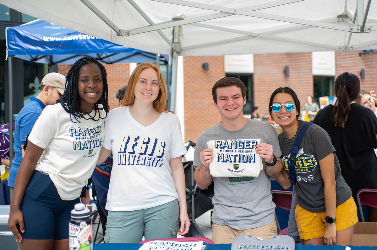 four students wearing Regis shirts smile for the camera under a tent on the Quad during Welcome Week at the Northwest Denver Campus